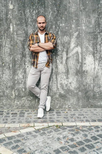 vertical shot of young good looking bold bearded guy standing outdoors against grey modern loft wall with his arms crossed and looking at the camera. wearing yellow shirt and jeans.