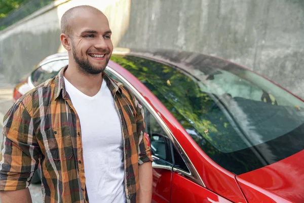 Młody dobry patrząc śmiały Brodaty facet stojący na zewnątrz w pobliżu jego czerwony samochód uśmiechnięty. noszenie żółtej koszulki — Zdjęcie stockowe