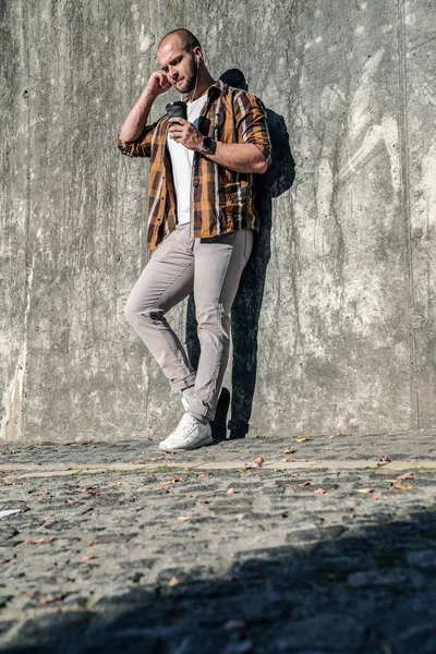 vertical shot of young good looking bold bearded guy standing outdoors against grey modern loft wall with cap of coffee in his hand and earphones listening music. wearing jeans and yellow shirt.