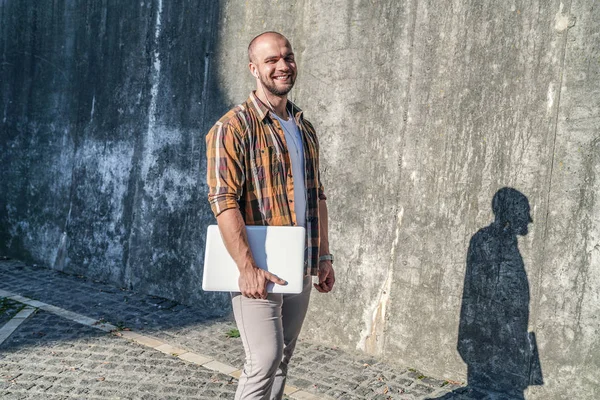 Good looking bold bearded freelancer standing outdoors against grey loft wall with his laptop and listens to music in earphones. looking at the camera and smiling. wearing yellow shirt and jeans.