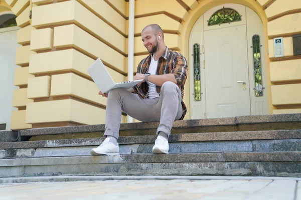 young good looking bold bearded guy is sitting outdoors on stairs in front of his house working on laptop. he is smiling and has earphones in his ears. wearing yellow shirt and jeans.