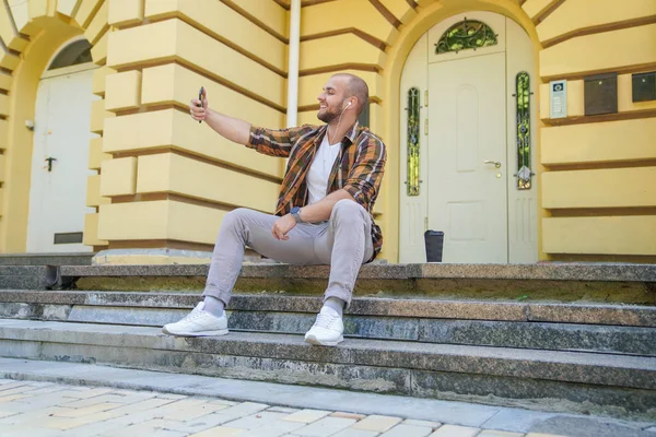 young good looking bold bearded guy is sitting outdoors on stairs in front of his house making selfie and smiling. he has earphones in his ears. wearing yellow shirt and jeans.