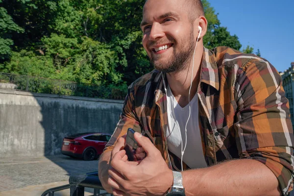 portrait of good looking bold bearded guy outdoors holding cellphone in his hand with earphones in his ears and listening music smiling.