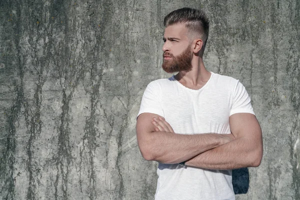 Side view portrait of young good looking bearded guy standing outdoors against grey modern loft wall looking aside with his hands crossed . wearing white shirt.