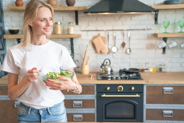Bela jovem sorrindo mulher segurando salada verde na cozinha. a olhar para o lado. Comida saudável. salada de legumes. Dieta. Estilo de vida saudável. cozinhar em casa. Vestindo camisa branca e jeans . — Fotografia de Stock