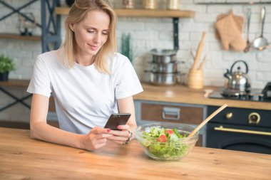 mutfakta salata yaparken cep telefonu kullanarak güzel genç kadın. Sağlıklı yiyecekler. Sebze salatası. Diyet. Sağlıklı yaşam tarzı. evde yemek pişirmek. Beyaz gömlek ve kot pantolon giymek.