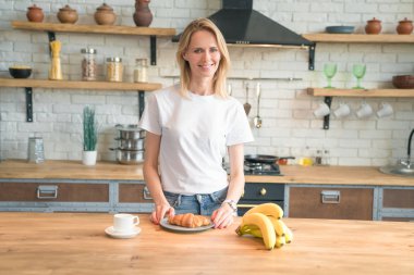 Güzel genç gülümseyen kadın evde mutfakta kahvaltı hazırlıyor sabah kahvesi, kruvasan, muz. Sağlıklı yiyecekler. Beyaz gömlek giymek