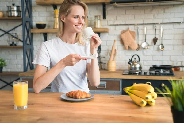 Jolie jeune femme souriante est assis dans la cuisine à la maison, prendre le petit déjeuner, boire du café avec des croissants et regarder de côté. Bonjour, Petit-déjeuner. portant une chemise blanche — Photo