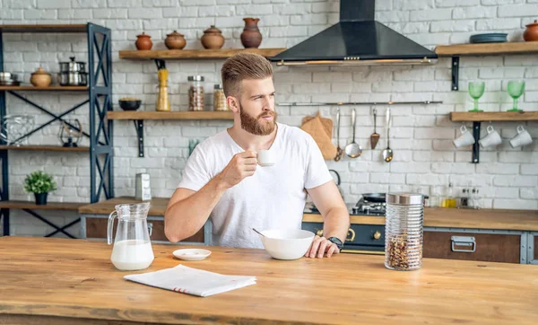 Beau beau barbu est assis dans la cuisine à boire du café du matin avec des céréales et du lait. lire la presse du matin, regarder de côté Alimentation saine. Mode de vie sain. portant une chemise blanche — Photo