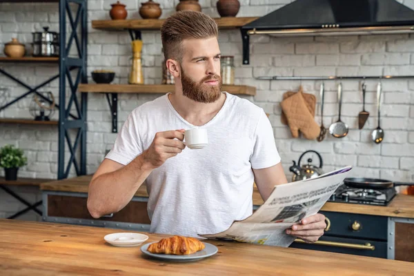 Good looking handsome bearded man is taking a rest sitting in the kitchen in the morning eating croissant with coffee, reading morning press and thinking, looking at the window. Healthy food.