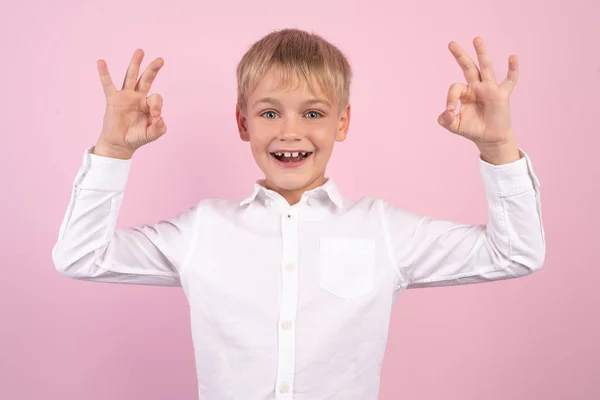 Портрет счастливого радостного маленького мальчика, улыбающегося и показывающего знак ОК обеими руками. студийный портрет на розовом фоне. в белой рубашке . — стоковое фото