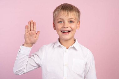 Beyaz gömlek giyen beyaz bir beyaz gülümseyen litlle çocuğun duygusal portresi, selamlaşmak için sağ elini geriyor. Mutlu çocuk el sallayarak, izole pembe arka plan.