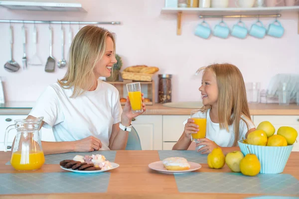 Zwei Blondinen, Mutter und Tochter, unterhalten sich in der Küche und trinken Orangensaft. Konzept für gesunde Ernährung. trägt weiße T-Shirts — Stockfoto