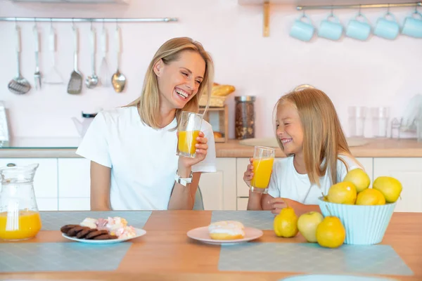 Zwei Blondinen, ein Profil von Mutter und Tochter lachen in der Küche und trinken Orangensaft. Frühstück mit Donuts und Schokoladenkeksen. trägt weiße T-Shirts — Stockfoto