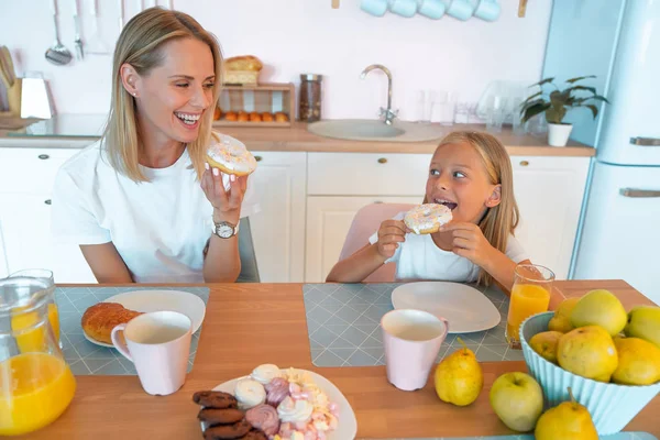 Mutter und Tochter essen leckere Donuts und haben Spaß miteinander. gekleidet in weißen T-Shirts auf farbigem Küchenhintergrund. — Stockfoto