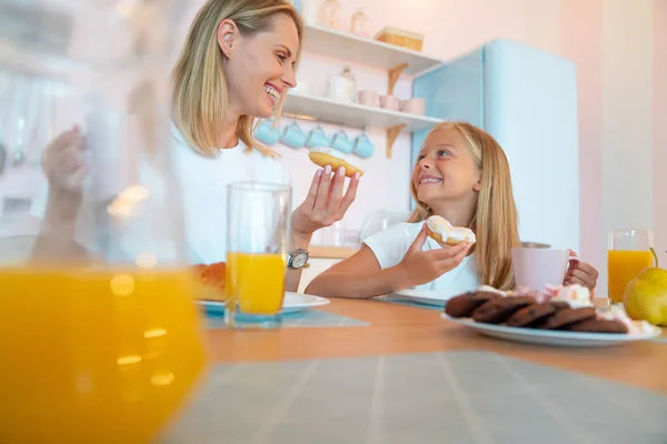 Mutter und Tochter essen leckere Donuts und haben Spaß miteinander. gekleidet in weißen T-Shirts auf farbigem Küchenhintergrund. — Stockfoto