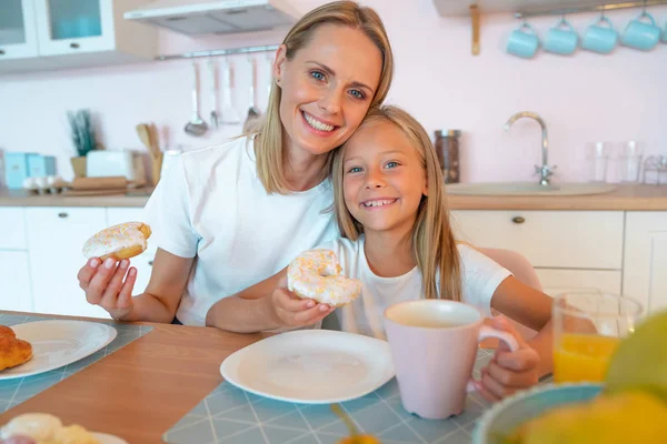 Linda foto de mamá y su hija desayunando con rosquillas y té. abrazar y mirar a la cámara —  Fotos de Stock