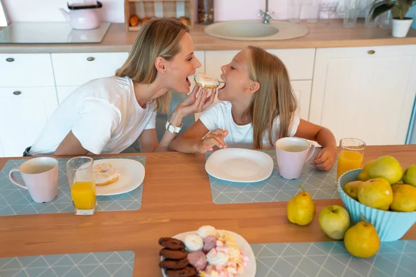Perfil de mamá e hija en la cocina mordiendo el donut en ambos lados. linda foto familiar. vestidos con camisetas blancas —  Fotos de Stock