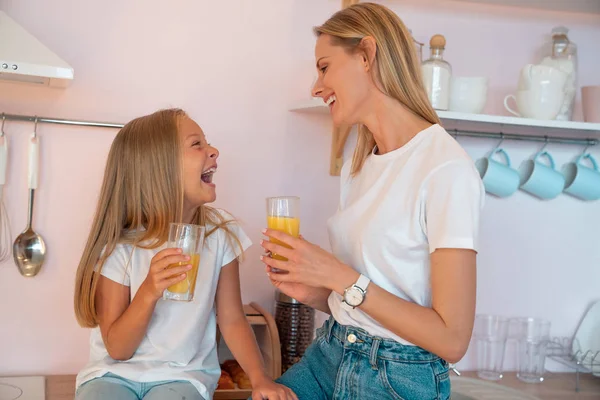 Feliz mamá bonita y su hija pequeña están bebiendo jugo de naranja en la cocina mirándose y riéndose. Familia feliz —  Fotos de Stock