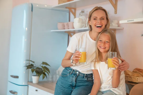 Mãe feliz e filhinha estão abraçando na cozinha em casa e bebendo suco de laranja sorrindo . — Fotografia de Stock