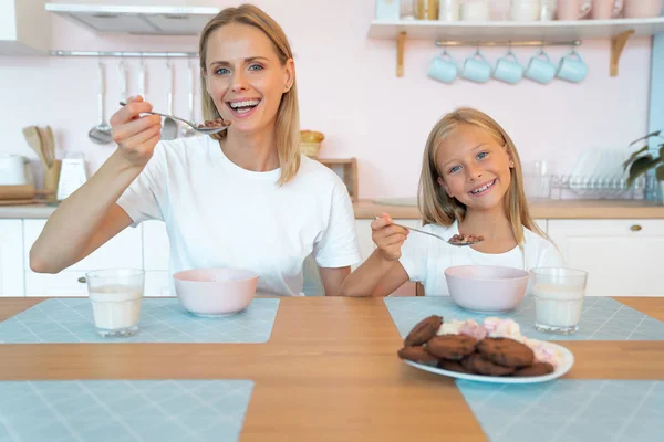 Mom with her daughter eating chocolate flakes. hold spoons by mouth and smile. have a good time together, dressed alike — Stock Photo, Image