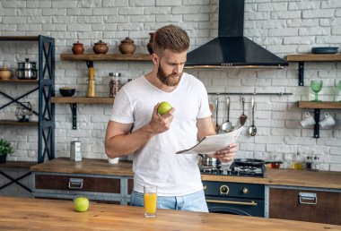 Yakışıklı sakallı adam elma yiyor ve yeni bir gazete okuyor. beyaz bir tshirt mutfakta duruyor