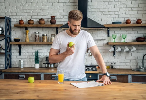 Beau barbu mangeant une pomme et lisant un journal frais. se tient dans la cuisine dans un t-shirt blanc — Photo