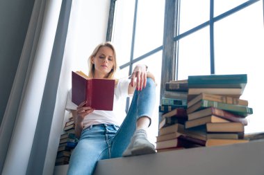 Düşünceli depresif, yalnız genç kadın fikir ve favori yazarın yeni bestseller arsa üzerinde kafa yoruyor şık coworking alanda rahat pencere üzerinde dinlenme.