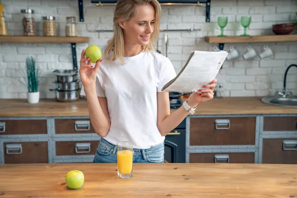 Jovem bela mulher está lendo sua imprensa da manhã e sorrindo enquanto drimking suco de laranja na cozinha. usando camisa branca . — Fotografia de Stock