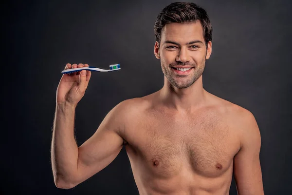 ハンサムな若いひげを生やした男が孤立した 手に歯ブラシや歯磨き粉を持ち笑顔で白髪の筋肉の男が灰色の背景に立っている 男性介護の概念 — ストック写真