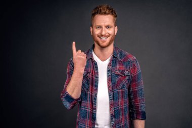 Bir numaralı jestinde işaret parmağı gösteren kıvırcık saçlı güzel saçlı, mutlu bir erkeğin kapalı çekimleri. Gri arka planda neşeyle gülüyor ve gülümsüyor..