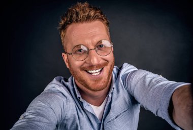 Kızıl saçlı adam internete yüklemek için selfie çekiyor. Tarz sahibi ve mutlu, sakallı ve şık saç kesimli yakışıklı bir adamın portresi. Kameraya doğru çekiliyor, sanki kendi fotoğrafını çekiyor, gülümsüyor gibi..