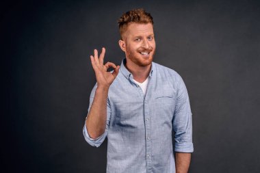 Mutlu iş ortağı işini kontrol altında tutuyor. Yakışıklı, sakallı ve kızıl saçlı olgun bir erkek. Gri arka planda poz verirken iyi ya da kötü bir jest yapıyor..