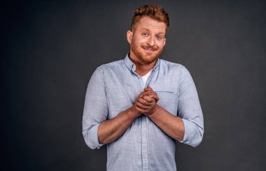 İyilik için yalvaran, yardım için minnettar olan mavi gömlekli, gri arka planda, sıkı ve umut dolu bir gülümseme içinde duran, avuçlarını göğsünün üzerinde tutan ve kaşlarını kaldıran, erkek giysisini bekleyen bir adam..