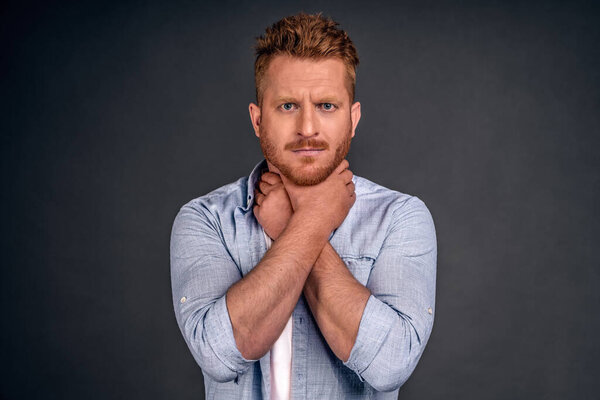 Unshaven displeased redhead man keeps hands on neck, suffers from suffocation, has sore throat, irritation, being unhealthy, looks unhappily at camera, stands against grey background, hard to swallow.