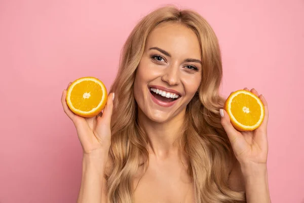 Attractive comic positive nude girl with beaming smile having two pieces of orange in hands, holding near face, isolated on pink background.