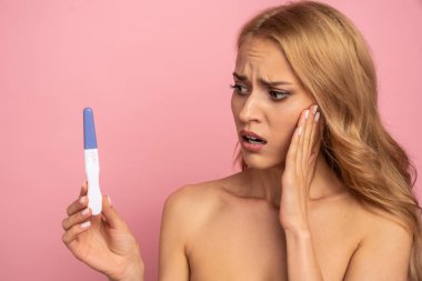 Aman Tanrım. Pembe arka planda izole edilmiş hamilelik testine bakarken hayal kırıklığına uğramış, beklenmedik üzgün bir modelin yüzü açık bir şekilde yüz yüze çekilmiş portresi..