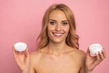 Çekici, genç, üstsüz, pembe arka planda izole bir şekilde duran, kavanozda yüz kremini gösteren sarışın kadının güzel portresi..