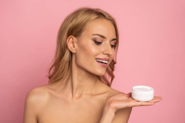 Neşeli modelin portresi ışıl ışıl gülümsemesiyle pembe arka planda izole edilmiş yağlanmış kuru cilt için etkili yüz maskesi ve beyaz kavanozu gösteriyor..