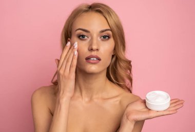 Güzel modelin portresi, etkili kremalı beyaz kavanozun olması, göz altına uygulanması, botoks etkisi, siyah halkalarla kavga etmek, pembe arka planda izole edilmiş kameraya bakmak..