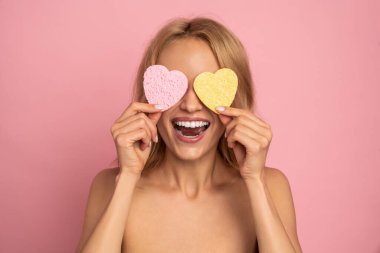 Aşırı neşeli komik kadının portresi kozmetik süngeri gözünün üstünde tutuyor, içtenlikle gülüyor, kırışıklıkları yok ediyor, cildini önemsiyor, doğal güzelliği var, pembe arka planda soyutlanıyor..