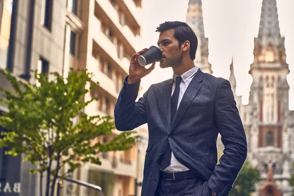 Coffee break. Confident young man in full suit holding coffee cup and looking away while standing outdoors with cityscape in the background