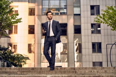 Kendine güveni tam genç ve yakışıklı bir adam, elinde bir fincan kahveyle arka planda şehir manzarasıyla merdivenlerde yürüyor.