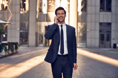 Kendine güvenen, takım elbiseli genç bir adam cep telefonuyla konuşuyor ve arka planda şehir manzarasıyla dışarıda dikilirken başka tarafa bakıyor.