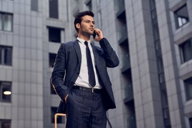 Kendine güvenen, takım elbiseli genç bir adam cep telefonuyla konuşuyor ve arka planda şehir manzarasıyla dışarıda dikilirken başka tarafa bakıyor.