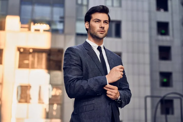 Confident businessman. Confident young man in full suit adjusting his sleeve and looking away while standing outdoors with cityscape in the background.