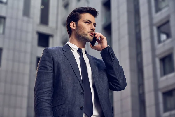 confident young man in full suit talking on the mobile phone and looking away while standing outdoors with cityscape in the background
