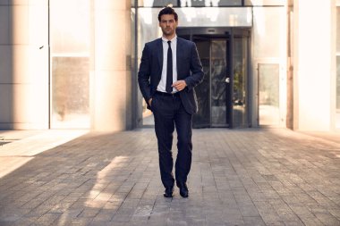 Genç ve başarılı. Yakışıklı genç işadamı arka planda ofis binasıyla dışarıda yürürken ceketini ayarlıyor ve başka tarafa bakıyor..