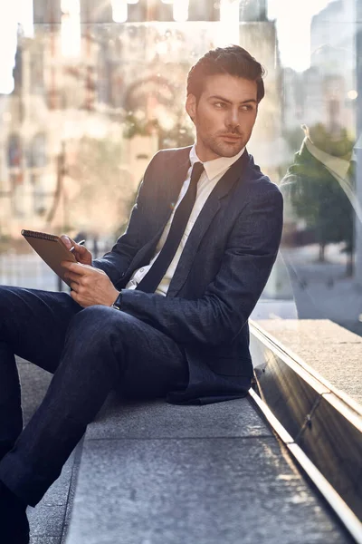 Confident happy young entrepreneur in formal suit writing in notebook while sitting at benche outdoors and looking away.