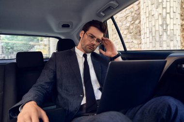 Ciddi ve düşünceli genç işadamı dizüstü bilgisayara bakıyor. Tek eliyle arabada otururken gözlük tutuyor..